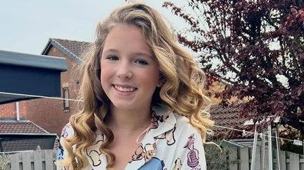An 11-year-old girl with long, curly, blonde hair stands in a garden. She is wearing a white shirt with pictures of cartoon characters on.