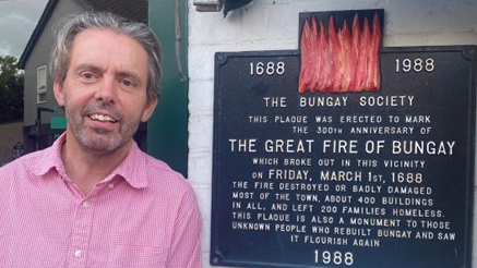 A man with swept-back grey hair and wearing a pink shirt stands next to a plaque which is mounted on a wall. A message on the plaque says it was erected to commemorate the 300th anniversary of The Great Fire of Bungay.