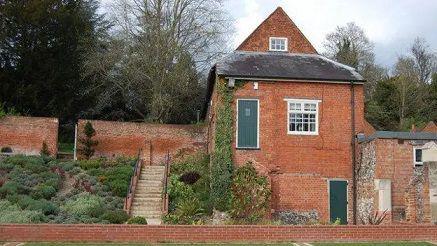 A red bricked Grade 2 listed former stables with slate roof, white windows, green doors and a sloped garden on the left with steps leading up to the top.   