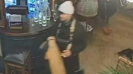 A blurred CCTV image of a man in a black outfit and beige hat inside a pub.