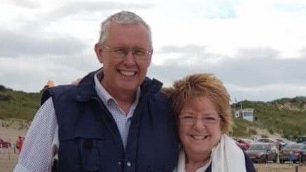 A couple with their arms around each other in front of sand dunes with cars in the background. The sky is cloudy and the couple are smiling. The man is wearing a navy gilet over a white shirt with grey short hair and glasses and the woman has ginger windswept spiky long hair with glasses and she is wearing a white scarf. 