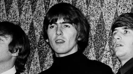 A black and white image of George Harrison. He has long shaggy hair and is wearing a black turtle-neck jumper.
