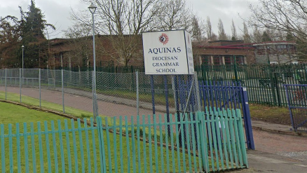 The front gates of a school, with a road travelling through the gates and into the background. Beside the gates is a sign reading Acquinas Diocesan Grammar School. In the background we can see a school building. 