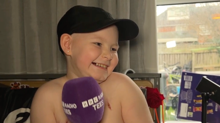 Riley is holding a purple Radio Tees microphone and is smiling broadly at the camera. The nine-year-old is wearing a black cap.