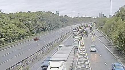 A CCTV image showing a line of traffic on a motorway, surrounded by trees. 