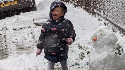 A young girl in boots and anorak stands in snow with a snowman.