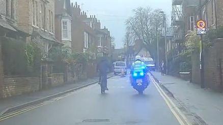 A police officer rides on a motorbike alongside a boy in dark clothing riding a push bike. They are rising along a residential street with a sign displaying a 20mph speed limit.