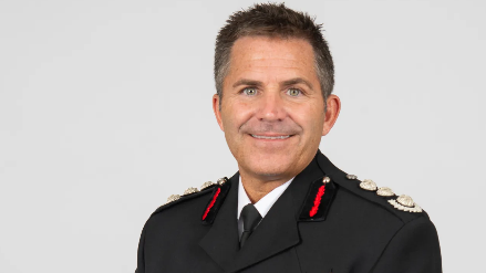 Simon Tuhill wearing his fire service uniform with a white shirt, black tie, black jacket and badges on his shoulders and lapel.