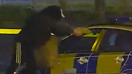 A figure in all black clothing points his arms out while holding an object directed at the window of a police car.
