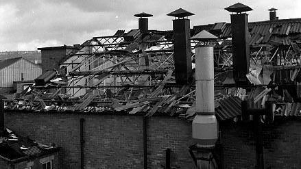 A black and white photo of the side wall and roof. Corrugated iron and debris are scattered around. At least half of the roof is missing