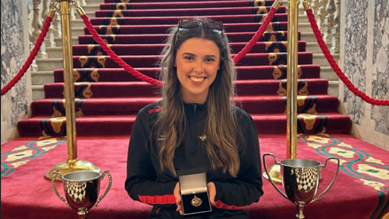 Méabh holding her medal. She's sitting on carpeted stairs with two sport cups beside her.