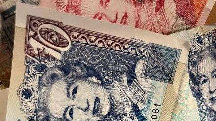 The face of Queen Elizabeth II can be seen on the corner of a £10 note. The note is dark blue and is lying on top of a red note. A third bank note can be seen to the right of this pile of money.