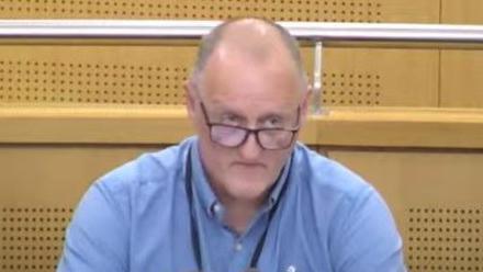 A man in a council chamber. He has plastic panels behind him and a metal railing. He is facing ahead, looking over the top of his glasses. He is balding and wears a blue shirt and black lanyard.