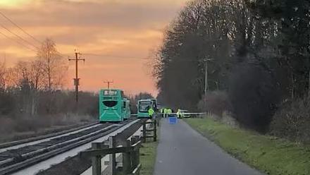 The guided busway between Cambridge and Histon with four green buses which have all stopped. Emergency service personnel in high-vis yellow jackets are on the scene. The footpath to the busway's right is closed. There are trees on both sides and the sky is pink and yellow at sunset.