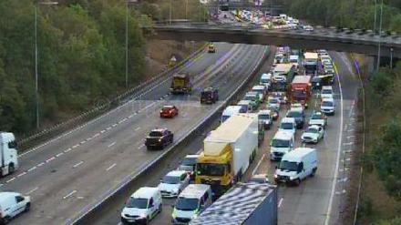 Long queue of cars on the M25 on one carriageway. The other carriageway is relatively clear. There is a bridge running across the road. 