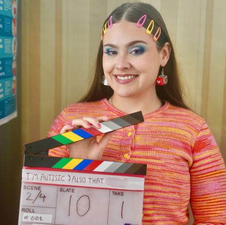 Molly is wearing a bright yellow, pink and red top. She has three hair clips each side of a centre parting in pink, yellow and orange. She's wearing red cube shaped earrings. She's holding a clapper board. She has bright blue eyeshadow and is smiling at the camera.  