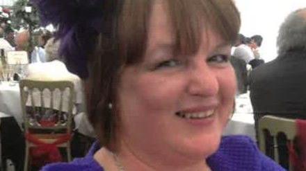 A woman dressed in a purple outfit with a matching headpiece, seated at a formal indoor event with other attendees in the background.