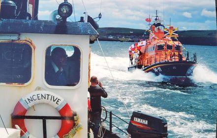 In the foreground is the cabin of the incentive with a life buoy on the side of it reading 'Incentive'. The boat is followed by a orange and blue RNLI boat