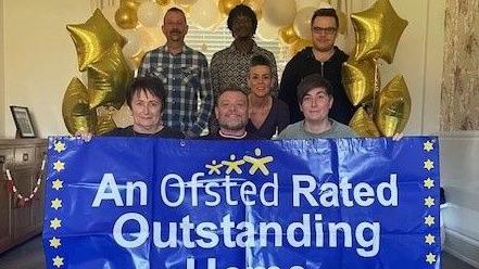 Four people three female and one male are crouching behind a banner stating "An Osfted Rated Outstanding Home" the banner is white writing on a blue background.  Three men are then standing behind them and under an arch of gold and white helium balloons, some of which are star shaped.