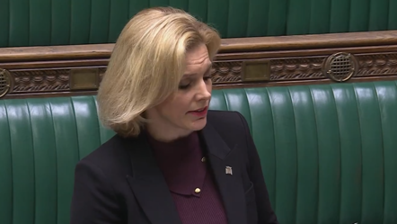 Woman infront of Parliament's green benches with short blonde hair purple top black blazer 