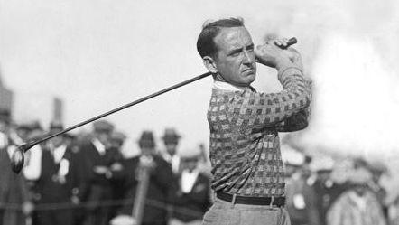 Old black and white image of golfer, Bobby Cruickshank, swinging golf club, with trousers tucked into socks, with crowd of smartly-dressed spectators in the background.