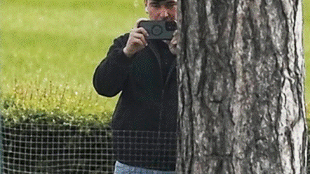 A man in a black coat stands behind a tree and a fence. He is partially obscured. The man is raising a smartphone and filming. 