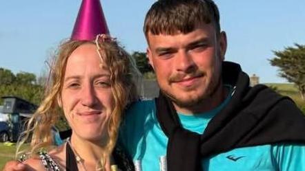 Jamie Chappell, a young man with dark hair and a beard, is standing in a field wearing a party hat next to a young woman holding a cake.