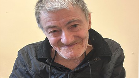 A head shot of an elderly man with grey hair and stubble, wearing a black quilted jacket.