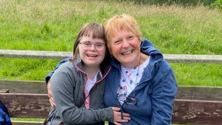 Elizabeth a girl with brown hair and a fringe wears glasses, she has a bright smile and sits next to her mum Frances, with short blonde hair in a coat, they sit on a bench in a field.