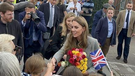 The Princess of Wales meets people outside a factory. She is shaking a member of the public's hand and is holding a large bunch of pink, red, orange and yellow flowers. People are waving Union flags. A group of men and women stand behind the princess and watch.