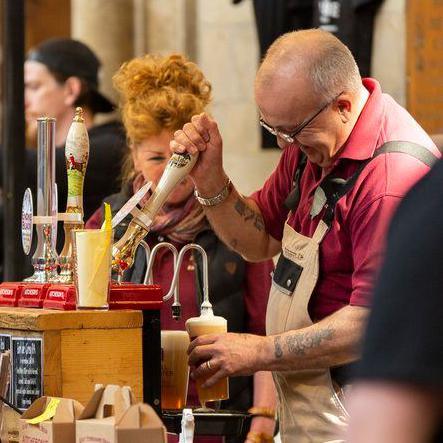 A man pulls a pint behind a stall at a beer festival, with a second pint resting. He wears a red shirt and cream apron. He is watched by a woman with red hair, whose face is hidden behind the beer pump.