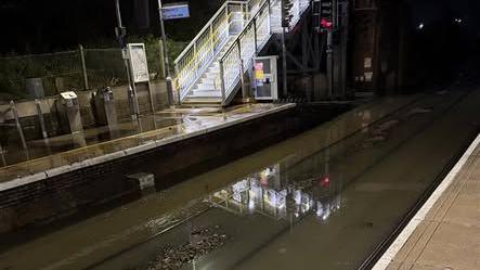 Entrance to a railway platform at night, with lights illuminating a stairway, and in the foreground is a waterlogged stretch of railway tracks. The water is dirty green and only the edge of the rails is visible.