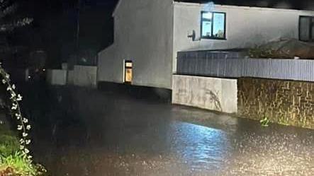 A flooded road next to a white house during the night time as heavy rain falls. A light is on in a window on the ground floor of the property and another is on in a larger window on the top floor.