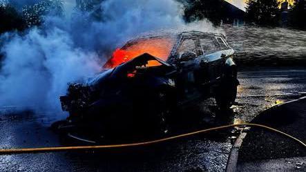 A black car on a road with bright orange flames inside it and white smoke emanating. A yellow hose can be seen crossing the road in front of it. It is sunset. 