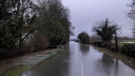 A flooded road with trees and countryside on the right-hand side and trees and large white house on the other side. 