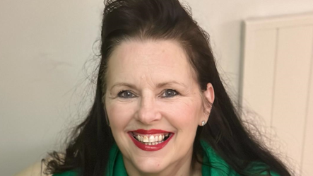 Shows woman in long dark hair, an earring in her left ear, red lipstick and a green hoodie smiling