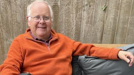 A grey-haired man wearing an orange fleece jumper with his arm spread across a sofa, wearing glasses