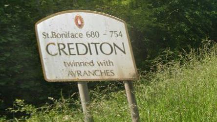 A road sign saying Crediton, St Boniface 680-754 twinned with Avranches on a grassy verge.