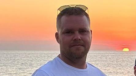 A man pictured wearing a t-shirt and sunglasses with a large body of water and a sunset in the background.
