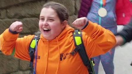 She has her brown hair in a ponytail and is walking and cheering, with her arms held up in victory. She is wearing a bright orange hoodie with the OSCARS charity branding.