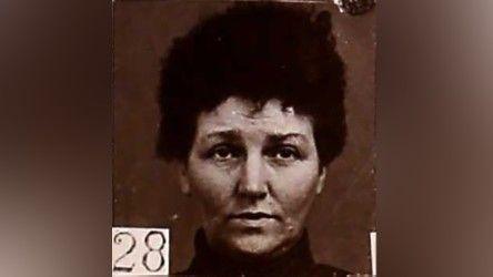 Black and white mugshot of a woman with dark hair piled on top of her head. She has a high collar around her neck and is looking slightly above the camera. There is a number - 28 - in the bottom left hand corner. 