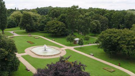Landscaped park with a central fountain surrounded by grass with flower beds. There is also paving, benches and trees.
