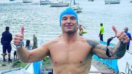 A man in multi-coloured swimming trunks sin front of a large body of water - posing with his thumbs up and wearing a blue swim cap.