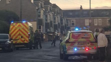 A residential street with a police car and ambulance which both have flashing blue lights. There are also spectators.