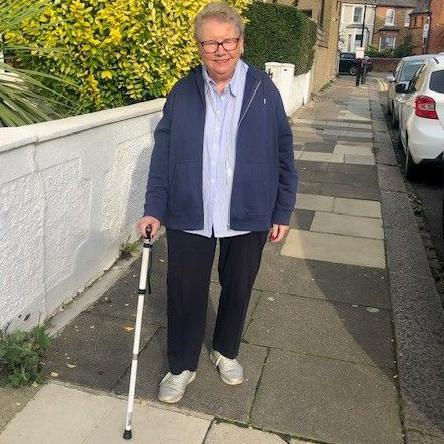 Hazel Kelly is walking along a street with the aid of a white walking stick. She is wearing glasses and a blue jacket and pale blue shirt and black trousers.