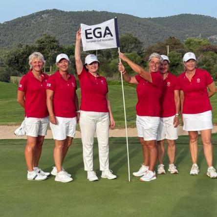 A group of six women stand together on a golf course, all wearing matching red shirts, white bottoms, and caps. One person holds a flag that says "European Golf Association". Behind them, green hills and trees stretch under a clear sky.