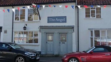 A building with a large light-teal coloured double front door with "The Angel" written above it. There are two cars parked in the street in front of it.
