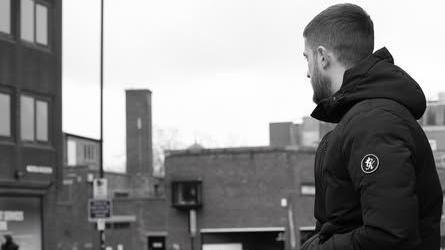 Man in winter jacket sits on a wall staring off-camera so his face cannot be seen. The photo is in black and white, and the mood is depressing.