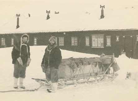 Two men stand on snow in front of a single-storey hut. They are wearing clothes for sub-zero conditions including hats and boots. They are standing in front of a large sledge.