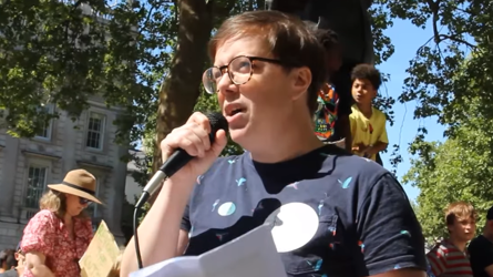 Clare from Berkshire is holding a microphone as she speaks to a crowd. She is wearing a blue T-shirt and has short, brown hair and is wearing tortoiseshell glasses.